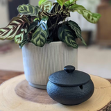 Black matte ceramic ashtray with lid placed on a wooden table beside a potted plant in a natural minimalist home setting