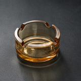 A clear amber-tinted glass cigarette ashtray with a thick base, photographed on a dark gray textured surface.