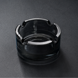 A dark gray smudge ashtray made of transparent glass, photographed on a dark gray textured surface.