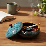 Ceramic ashtray with blue lid on a wooden table with a smoking cigarette inside.