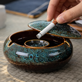 Close-up of large ceramic ashtray with lid and cigarette rests, hand placing cigarette into glossy blue patio ashtray